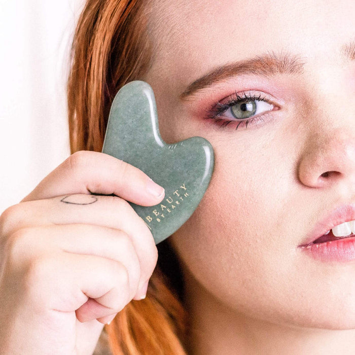 A person uses a Beauty by Earth Jade Gua Sha on their cheek, enhancing their beauty ritual. Their pink eyeshadow complements light skin and reddish-brown hair. A small hand tattoo is visible as they gracefully hold the tool, which bears the BEAUTY etching.