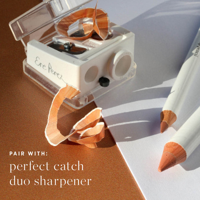 A white dual sharpener with wood shavings next to two sharpened Ere Perez Jojoba Eye Pencils on a white surface. Text: Pair with Perfect Catch Duo Sharpener—ideal for sensitive eyes, enriched with jojoba.