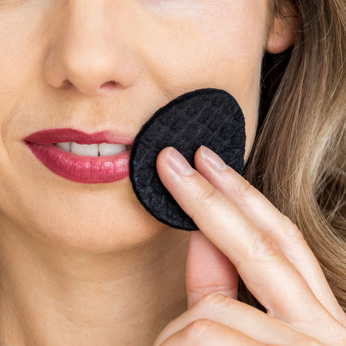 A person gently removes makeup from their cheek with the eco-friendly LastRound Pro by LastObject, highlighting the reusable cotton round and their striking features against a backdrop of pink lipstick.