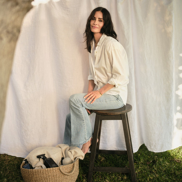 A woman with long dark hair sits barefoot outdoors on a wooden stool. Beside her, a woven basket holds Homecourt Linen Spray and an eco-friendly beige cloth. A white sheet hangs in the background.