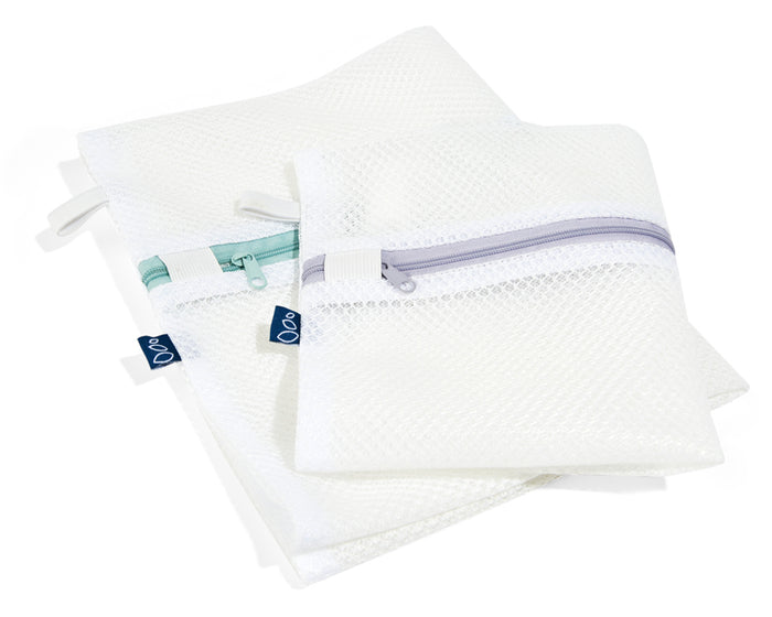 Two overlapping Grove Co. Laundry Delicates Bags, ideal for delicates, rest on a white surface; the larger bag has a green zipper, and the smaller one has a gray zipper. Both reusable bags feature small black tags.
