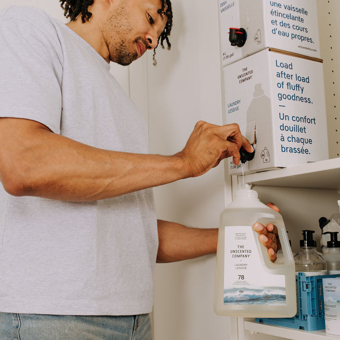 In a tidy laundry area, someone refills a large plastic bottle with Laundry Detergent Refill from The Unscented Company’s box dispenser.