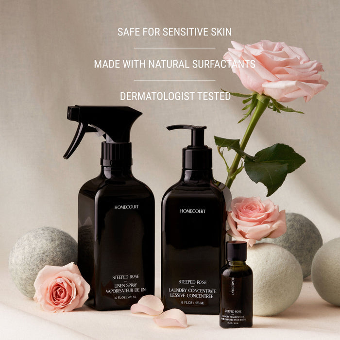 A black Homecourt Linen Spray bottle sits with pink roses and gray stones. Text above reads: Safe for sensitive skin, eco-friendly, made with natural surfactants, dermatologist tested.