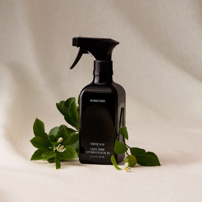 A black Homecourt Linen Spray bottle rests on a light fabric, surrounded by green leaves and white flowers, emphasizing its signature scent made with eco-friendly ingredients.