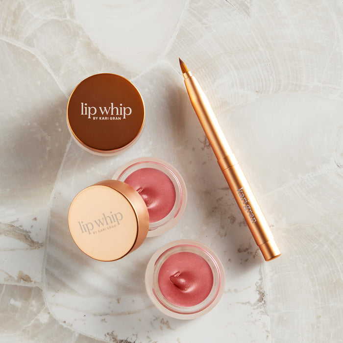 Two small open jars of Kari Grans Lip Whip - Peppermint Blush, paired with a matching lip brush featuring a metallic cap, sit elegantly on a marbled surface. A closed jar with a gold lid is nearby. This organic lip care product epitomizes luxury and pampering.