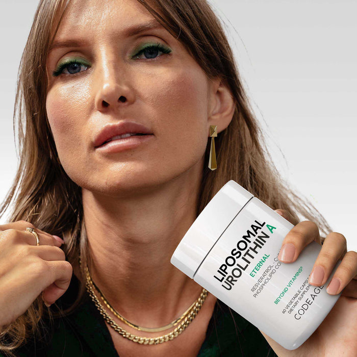 A woman with light brown hair and green eyeshadow confidently holds Codeage Liposomal Urolithin A close to her face, highlighting its benefits. She wears gold jewelry and poses against a plain background.