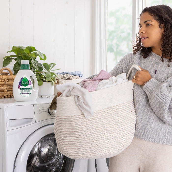 A woman in a gray sweater stands in a bright laundry room with a basket, beside her are a washing machine, potted plant, and a bottle of Seventh Generation Liquid Laundry Detergent, known for its natural ingredients. She looks to the side, embracing eco-friendly choices.