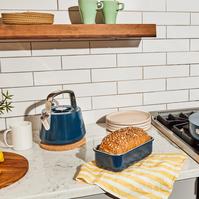 A kitchen features a blue kettle on a marble countertop, with bread in a non-toxic Caraway Loaf Pan on a yellow-striped cloth. A wooden shelf displays green mugs against white subway tiles.