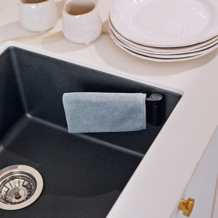 A modern kitchen sink with a gray sponge holder and a HAPPY SiNKS Magnetic Dishcloth Holder attached. Nearby, white ceramic cups and marble-patterned plates are on the counter. The sink includes a metallic drain featuring an innovative Magnet Fitting System.