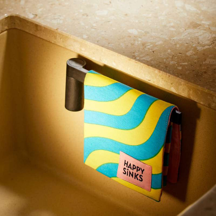 A Magnetic Dishcloth Holder by HAPPY SiNKS in a yellow and blue pattern is elegantly draped over a black hook next to a beige sink, highlighting its biocomposite material and Magnet Fitting System for secure placement.