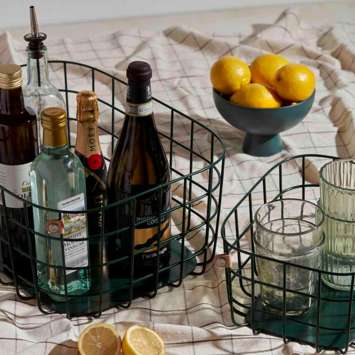 Decorative Open Spaces Medium Wire Baskets hold wine and spirit bottles on a checkered tablecloth. A bowl of lemons sits nearby, with more lemons scattered around. Tall glasses are in another stylish basket made from high-quality painted steel for an extra touch of flair.