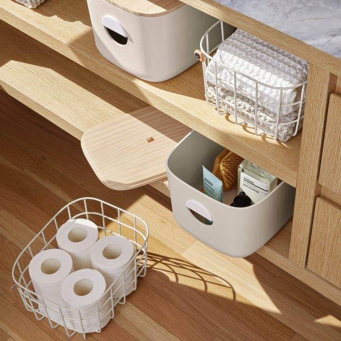 Adorned with Open Spaces Medium Wire Baskets, a wooden shelving unit showcases a minimalist bathroom setup—one basket holds rolled white towels, another displays toilet paper rolls, while the third organizes toiletries for a high-quality, organized aesthetic.