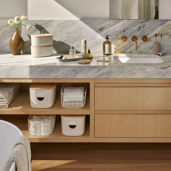 A bathroom vanity with a marble countertop displays a modern sink and brass fixtures. It holds Open Spaces Medium Wire Baskets for toiletries, alongside a floral vase. Natural light highlights the wooden cabinetry and sleek decor, ensuring seamless organization.