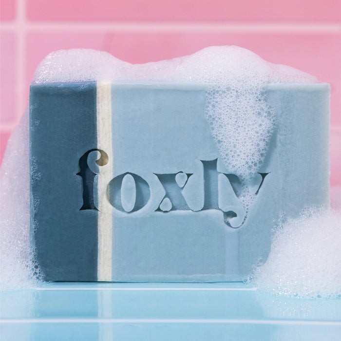 Foxly Moisturizing Bar Soap, a blue and white handcrafted bar with "Foxly" etched on it, rests foamy on a wet surface before pink tiles—crafted from clean ingredients and nourishing oils for a gentle cleanse.