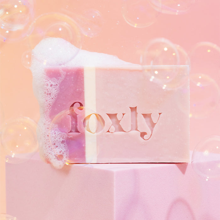 A Foxly Moisturizing Bar Soap sits on a pink surface, covered in foam and bubbles against a soft background, highlighting its clean ingredients and nourishing oils.