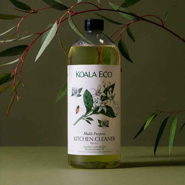 A bottle of Koala Eco Multi-Purpose Kitchen Cleaner Refill sits on a green surface with leafy branches behind it. The label features botanical illustrations and emphasizes the cleaner’s natural ingredients.