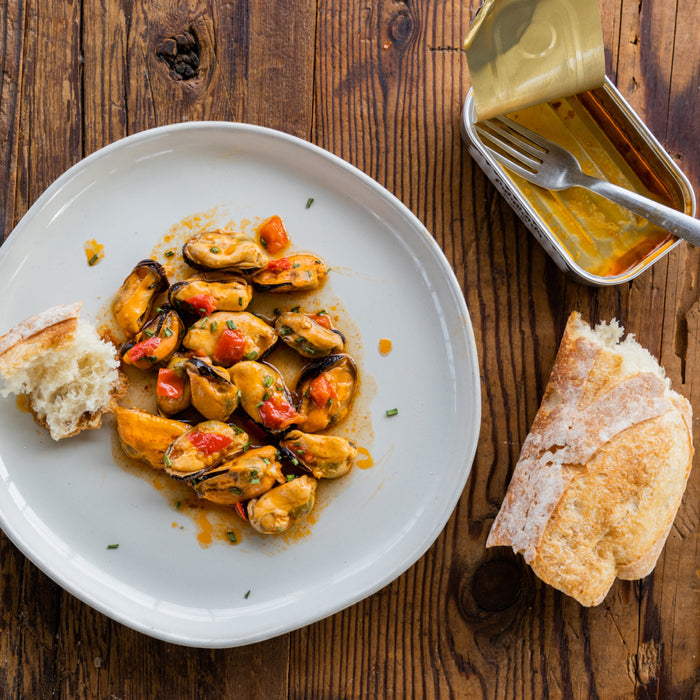 A plate of Patagonia Provisions Mussels Spicy in sauce with diced red peppers and rustic bread, drizzled with extra virgin olive oil, sits on a wooden table. An opened tin of mussels and an extra piece of bread are beside the plate.
