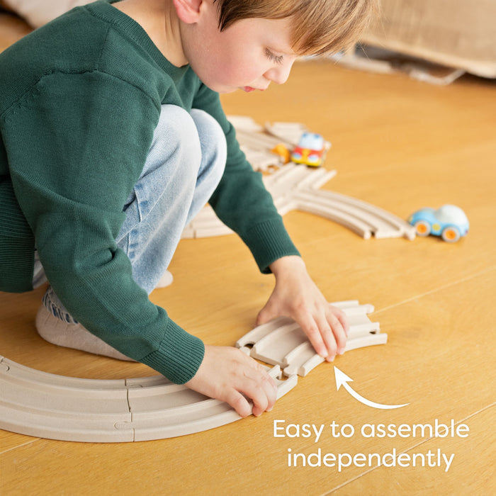 A young child in a green sweater and jeans kneels on the floor assembling HABAs My First Racetrack – made from 100% recycled materials for ages 2+. Two cars are on the track. Text reads: Easy to assemble independently.