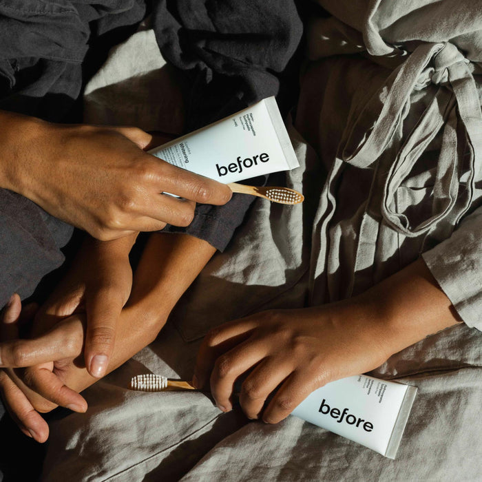 Two people wearing neutral clothing lie side by side, gently touching hands while holding before n-HA Whitening Toothpaste Fluoride-Free and bamboo toothbrushes.