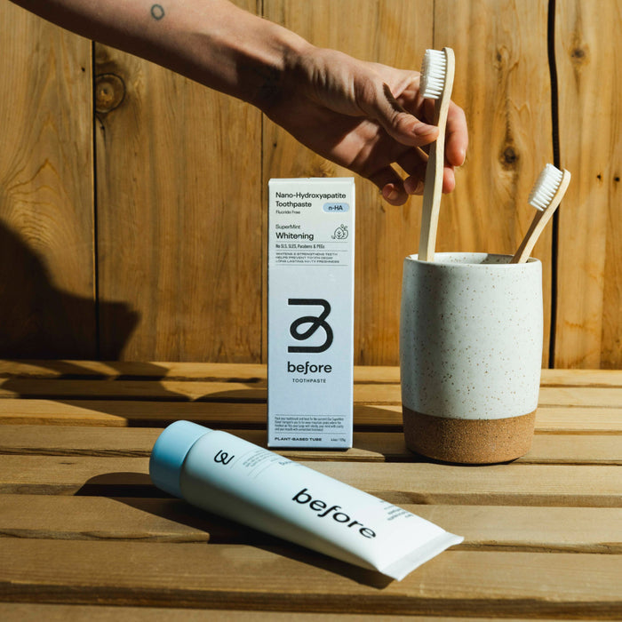 A hand places a bamboo toothbrush into a speckled ceramic cup holding another toothbrush. Nearby, before n-HA Whitening Toothpaste Fluoride-Free by Before sits on a wooden surface with its box, set against a wood background.