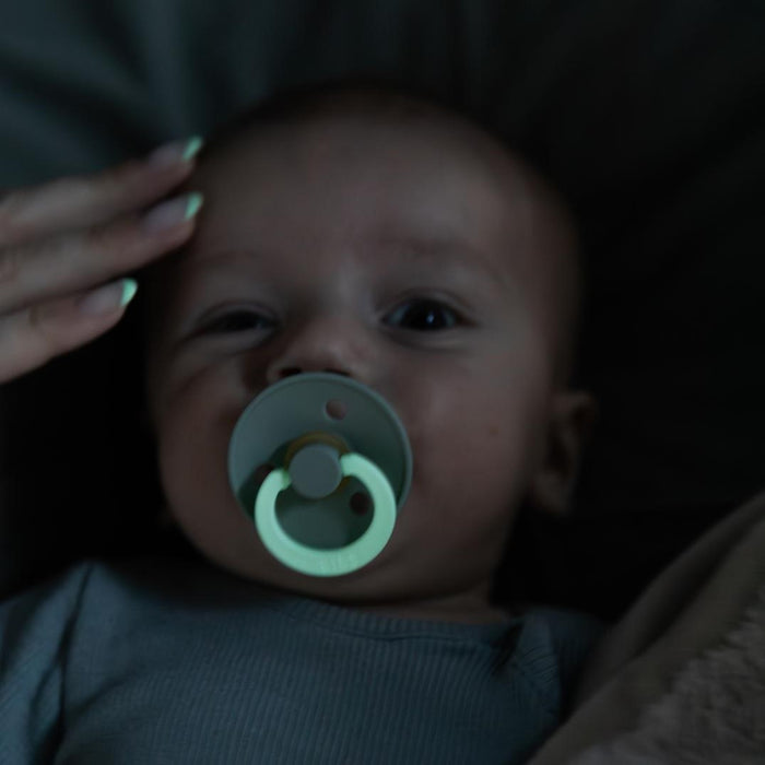 A baby peacefully lies on a dark surface with a BIBS Natural Latex Rubber Glow in the Dark Pacifier, Size 2 (6-18 Months) in their mouth, softly illuminated by dim light as a hand tenderly rests on the babys forehead.