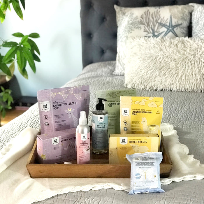 A wooden tray filled with eco-friendly laundry items, including detergent pods and stain remover spray, rests on a neatly made bed with light gray bedding. Newborn Baby Dryer Sheets - Chamomile by grab green promise to soften fabrics and fight static cling, while decor like a plant and starfish cushion adds charm.