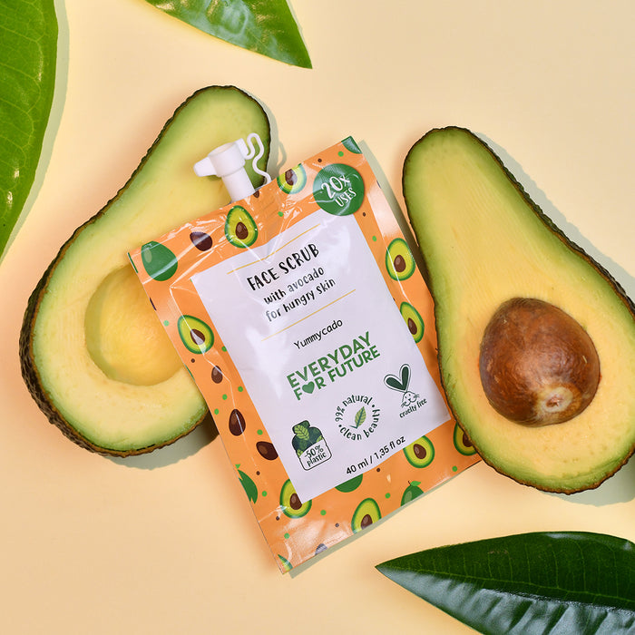 The Nourishing Avocado Face Scrub packet by Everyday For Future, featuring natural ingredients and adorned with avocado illustrations and sustainability icons, is nestled between two avocado halves on a light yellow background. Green leaves partially frame this eco-friendly skincare essential.