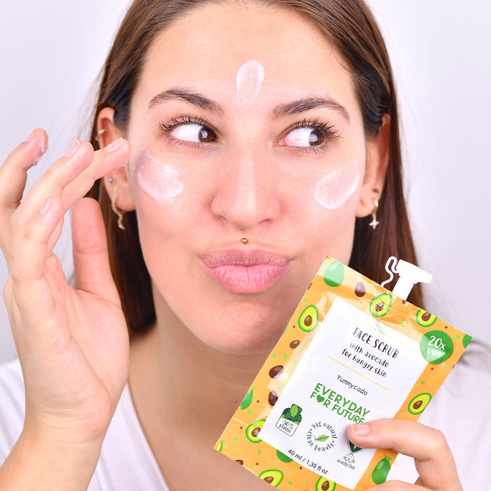 A woman playfully applies Everyday For Futures Nourishing Avocado Face Scrub from a pouch with avocado images, leaving dots on her cheeks and forehead, hinting at a fun skincare routine enriched with natural ingredients for flawless skin.