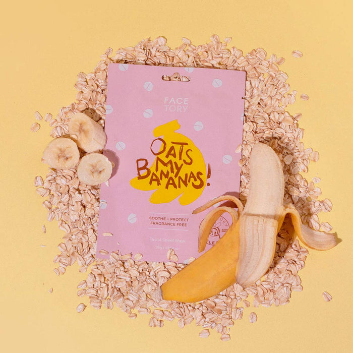 A pink package of FaceTorys Oats My Bananas Sheet CALMING Mask rests on scattered oats, surrounded by banana slices and a peeled banana on a light yellow background. This soothing formula uses oat extract for hydration.