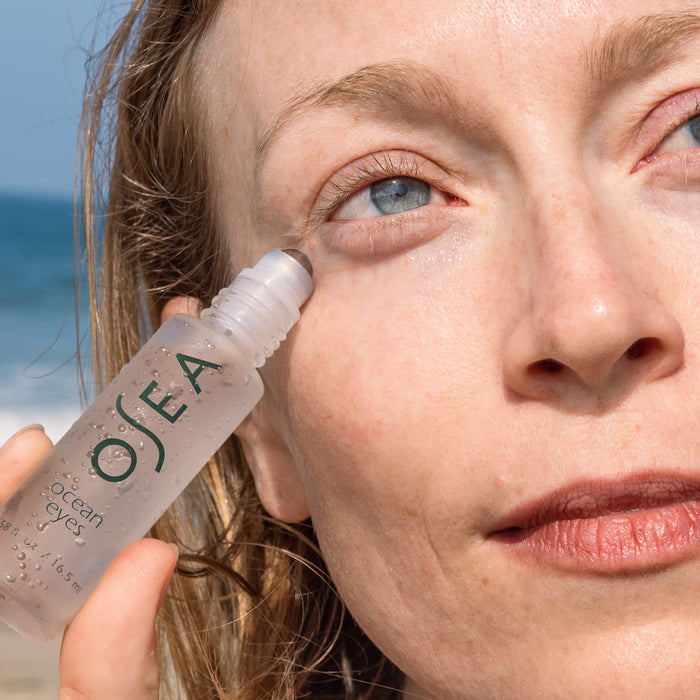 A person uses OSEAs Ocean Eyes® Age-Defying Eye Serum near their eye to tackle dark circles. With wet hair and an upward gaze, they find tranquility and embrace self-care by the sea as a blurred beach and ocean backdrop offer serenity.