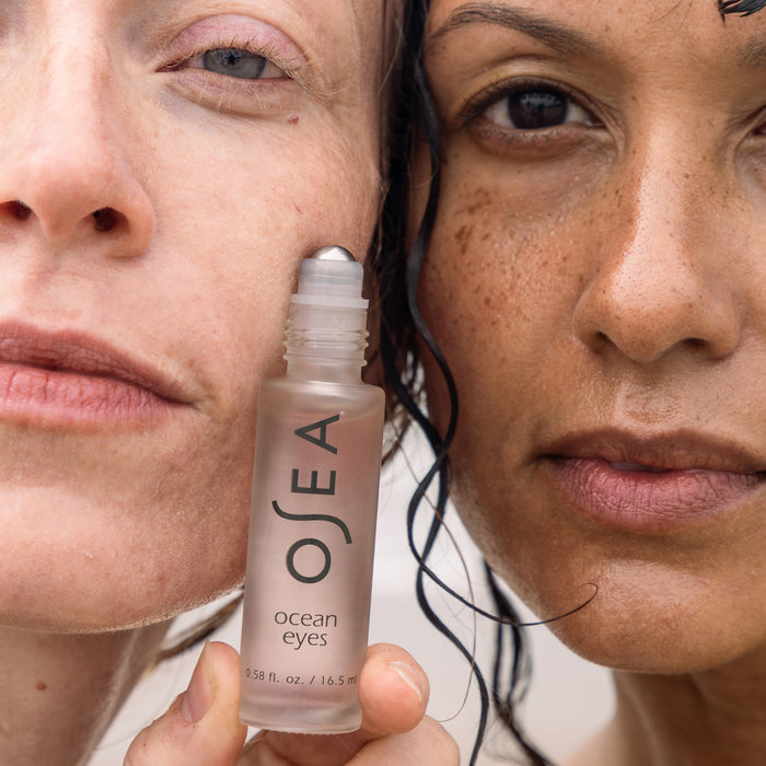 Two people hold a clear bottle labeled OSEA Ocean Eyes® Age-Defying Eye Serum with close-up facial expressions. The hydrating gel serum, featuring a roller applicator, enhances their facial features and promises to reduce dark circles while rejuvenating the skin around their eyes.