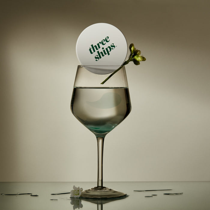 A glass of clear liquid sits on a reflective surface, topped with a Three Ships coaster. A small yellow-green flower rests nearby, evoking the refreshing benefits of Off Duty Sea Algae + Bio-Peptide Smoothing Eye Masks by Three Ships.