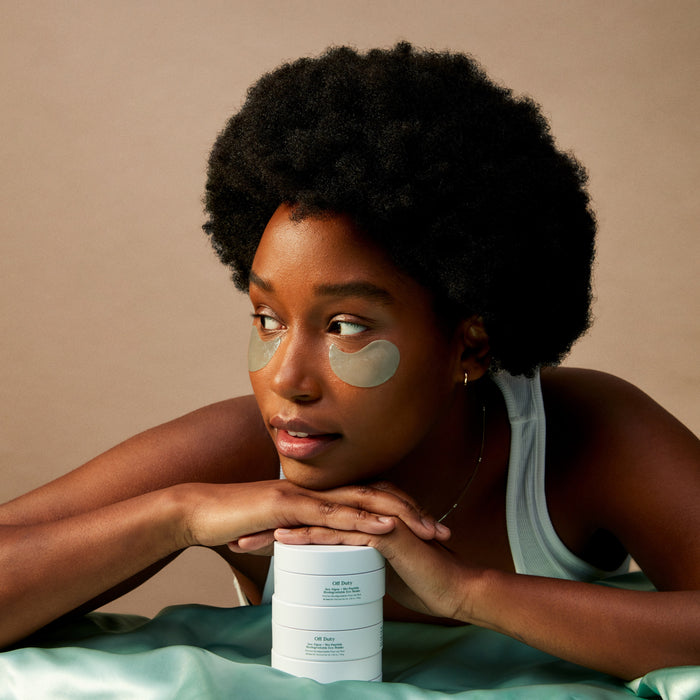 A woman with natural curly hair, in a white tank top, rests her chin on skincare jars while wearing Three Ships Off Duty Sea Algae + Bio-Peptide Smoothing Eye Masks, looking to the side against a neutral background.
