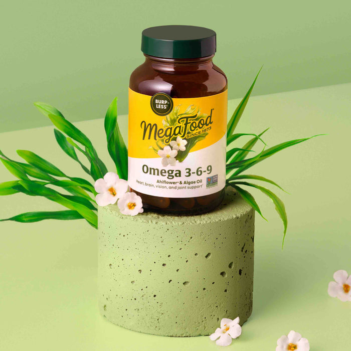 A bottle of MegaFoods Omega 3-6-9 dietary supplement, adorned with a yellow label, sits on a green cylindrical stand. The scene is completed with decorative green leaves and small white flowers, all set against a light green backdrop.