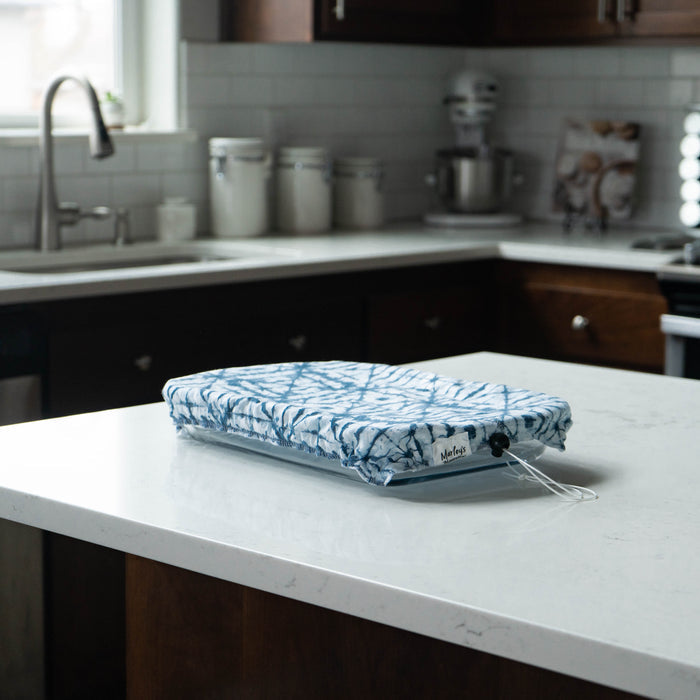A blue and white patterned fabric bag sits on a kitchen countertop with subway tiles and dark cabinets. Nearby appliances and three white canisters are near the reusable Marleys Monsters Pan Cover Bundle made from food-safe PUL, tucked inside.