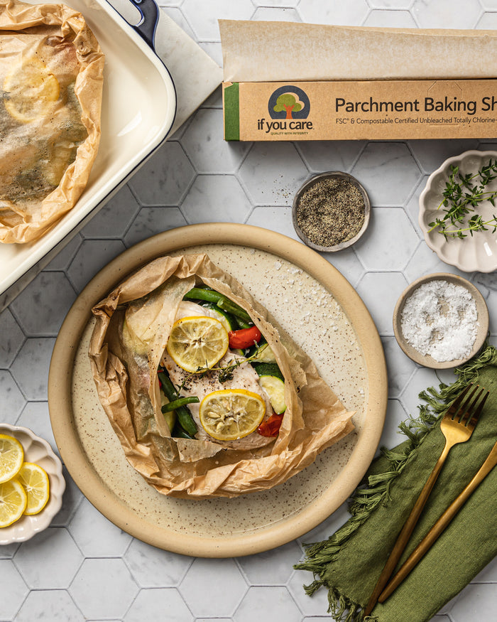 Parchment paper-wrapped chicken with lemons, tomatoes, and green beans is served. A box of If You Care Parchment Baking Sheets, salt, pepper, lemon slices, and gold utensils sit on a green napkin on the tiled surface.