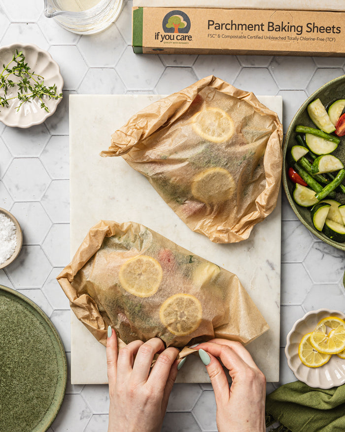 Two hands wrap food parcels with lemon slices in If You Care Parchment Baking Sheets on a marble surface. Nearby are a box of these sheets, herbs on a small plate, seasoned veggies in a dish, and a green plate. Lemon slices rest on a small wooden board.