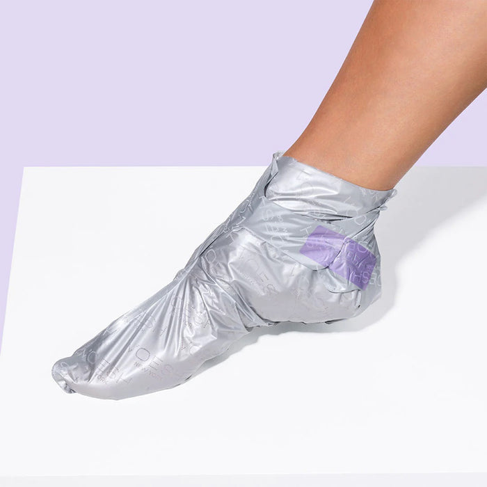 A foot enclosed in a shiny gray VOESH New York Peeling Socks Duo rests on a white surface against a light purple background.