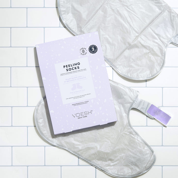 A package of VOESH New Yorks Peeling Socks Duo, noted for vegan ingredients, is displayed on a white tiled surface. One sock peeks out from the light purple box with a lavender tab, featuring text and the logo.