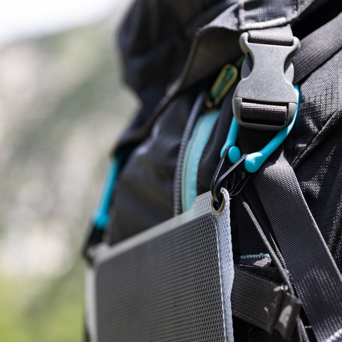 Close-up of a backpack strap with a plastic buckle, blue zipper, and carabiner clip—perfect for attaching the Paleblue Portable Solar Charger (7 Watts Max Output) for portable energy; blurred green and white outdoor scenery in the background.
