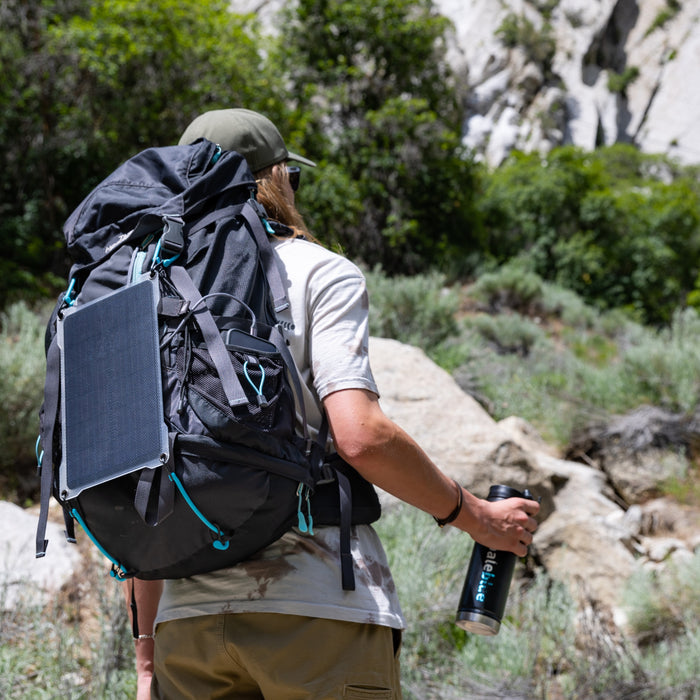 A hiker with a large backpack and Paleblue Portable Solar Charger (7 Watts Max Output) attached treks a rocky, green trail, carrying a water bottle and wearing a cap with a light shirt.