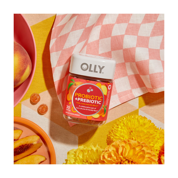 A jar of OLLY Probiotic + Prebiotic is elegantly displayed on a checkered cloth with sliced peaches and orange gummies. Yellow flowers and a pink dish complete the vibrant scene, creating an appealing visual for those focused on digestive health.