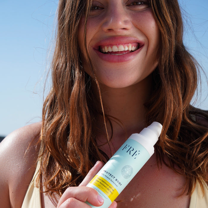 A smiling woman with long brown hair enjoys the sun outdoors, holding a bottle of FRÉ Skincare PROTECT BODY 100% Mineral Sunscreen SPF 50 for powerful broad spectrum protection under the clear blue sky.