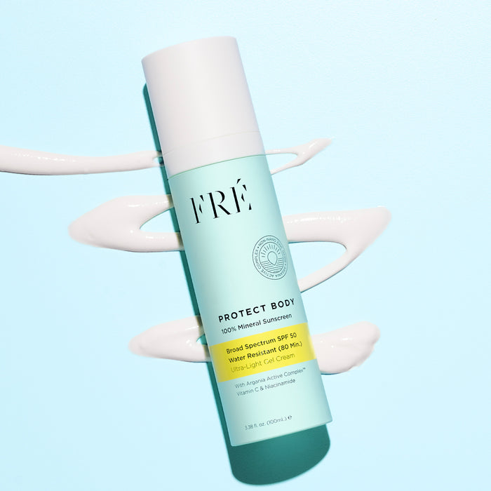 A turquoise bottle of FRÉ Skincare PROTECT BODY 100% Mineral Sunscreen SPF 50 lies diagonally on a pale blue background, with white sunscreen swirls beneath it to highlight its broad spectrum protection.