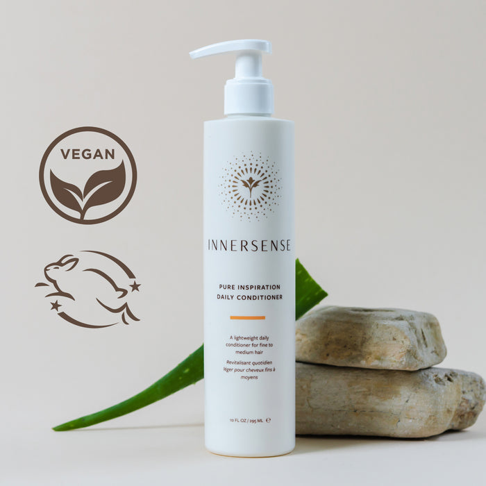 A bottle of Innersense Pure Inspiration Daily Conditioner is placed against a neutral background, accompanied by a sliced aloe leaf and a rock. The vegan and cruelty-free logos are visible, indicating product attributes.