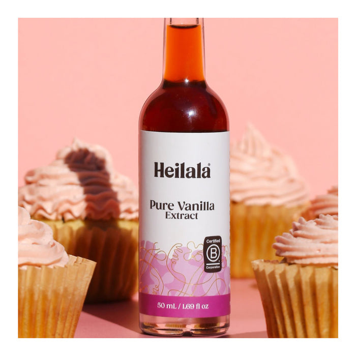 A bottle of Heilala Pure Vanilla Extracount sits on a pink surface among cupcakes with pink frosting, highlighting the rich, ethical vanilla flavor against a soft pink background.