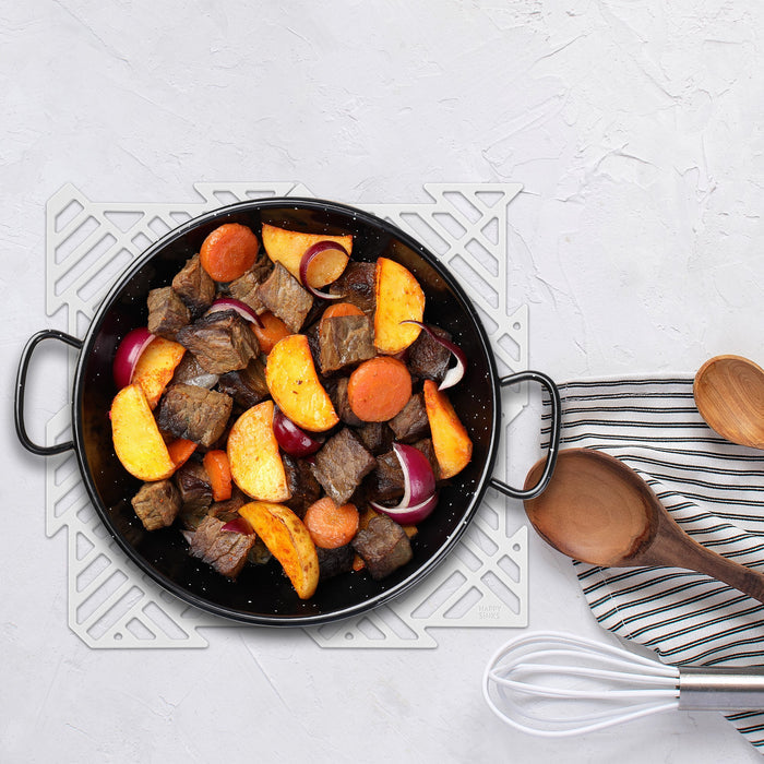 A decorative trivet holds a black pan with cooked beef, sweet potatoes, onions, and carrots. Nearby, utensils made from food-grade silicone sit neatly on a striped cloth over the tidy kitchen’s white textured surface with a Puzzle Sink Mat by HAPPY SiNKS.