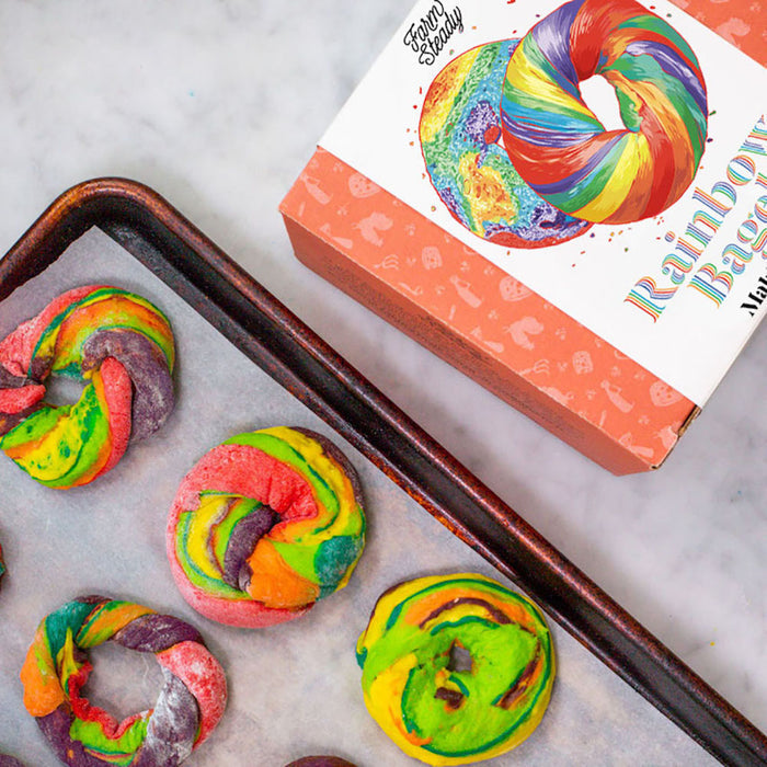 A Rainbow Bagel Making Kit from FarmSteady, featuring a tray of vibrant rainbow bagels with hues like purple, green, yellow, and red, sits next to a box displaying similarities. Perfect for DIY enthusiasts eager to create colorful homemade bagels.