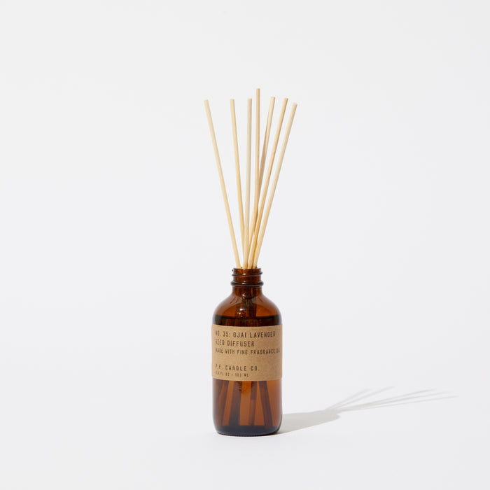 A brown glass Reed Diffuser by P.F. Candle Co., labeled Lavender with teakwood notes, stands elegantly on a white surface with wooden reeds inserted. Its shadow extends rightward, enhancing the minimalist scenes depth and charm.