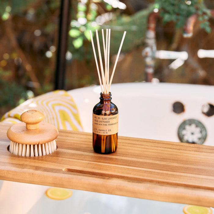 A wooden bath tray holds a P.F. Candle Co. Reed Diffuser with reeds, exuding lavender notes, and a bristle brush with a wooden handle. A white jet bathtub with teak accents is next to green plants outside the window.